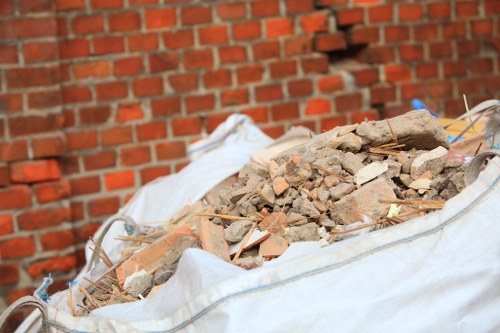 A large white sack filled with a pile of mixed construction debris, including broken bricks, concrete fragments, and small pieces of wood, is positioned outdoors on a paved surface. The debris is predominantly reddish-brown, with some grey cement pieces and lighter wood fragments visible. The sack appears to be made of woven polypropylene material, and the contents are uneven and loosely stacked. In the background, there is a red brick wall with a typical bond pattern, providing an urban or residential setting context. The lighting suggests natural daylight, illuminating the debris clearly, with some shadows cast across the scene. This image represents a typical scene of rubbish removal involving building waste, suitable for a company like Commercial Waste Removal Leyton, which offers rubbish clearance services in the Leyton area, within the E11 postal code zone.