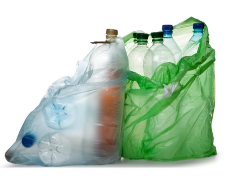 A collection of several used plastic bottles and containers placed inside a partially open green plastic rubbish bag, with some bottles showing indentations and labels, set against a plain white background. The bottles vary in size and colour, with clear, green, and blue hues, and are made of PET plastic with smooth, shiny surfaces. The green rubbish bag is crumpled around the bottles, revealing the tops and necks of the bottles at the opening, and appears to be lightweight and flexible. The scene is well-lit, emphasizing the transparency and textures of the plastic bottles and the glossy finish of the bag. This image relates to waste management services, such as those offered by Commercial Waste Removal Leyton, on a typical disposal or recycling task. The focus is on the visual details of household plastic waste ready for collection or recycling, set in a neutral context suitable for a waste removal website serving Leyton or nearby areas.