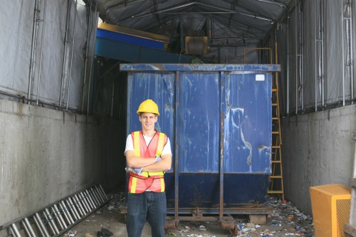 A young male waste management worker stands confidently in front of a large, blue, industrial skip filled with mixed rubbish inside a covered loading area. He is wearing a yellow safety helmet, a high-visibility orange vest with reflective stripes, a white shirt, and dark work trousers. The skip is situated on a concrete floor, surrounded by scattered debris and waste materials, including paper and plastic, indicating recent rubbish collection or removal. To the left, a metal ladder leans against the wall, while on the right side, a yellow waste bin is partially visible. The environment appears industrial, with steel walls and a curved, metallic roof overhead, suggesting an internal loading or disposal site typical in commercial waste removal services in Leyton or nearby areas. The overall scene captures a professional waste collection operation managed by Commercial Waste Removal Leyton, emphasizing efficient rubbish clearance within an urban setting, consistent with local waste management requirements.