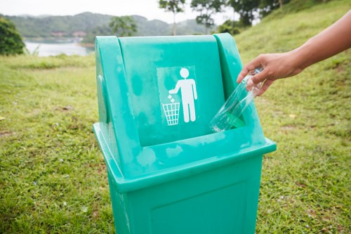 A person's hand is in the process of discarding a clear plastic bottle into a bright turquoise recycling bin situated outdoors on a grassy area. The bin features a white symbol of a human figure disposing of rubbish into a bin, indicating its purpose for recyclable waste. The background reveals a scenic view of a lake or river, with lush green trees and distant hills, suggesting a park or natural environment near Leyton or the surrounding postcode area. The scene is well-lit, highlighting the texture of the plastic bottle and the smooth, clean surface of the bin. The grassy ground beneath the bin is evenly mowed, providing a tidy setting for waste disposal. This image demonstrates appropriate rubbish disposal behavior, aligning with services provided by Commercial Waste Removal Leyton for maintaining clean outdoor spaces in the local community.